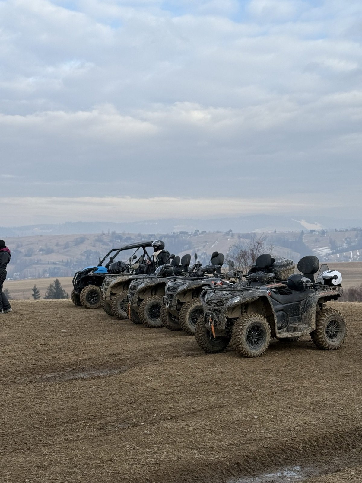 Quady i buggy ustawione w rzędzie na zaoranym polu, w tle pasmo gór pod zachmurzonym niebem