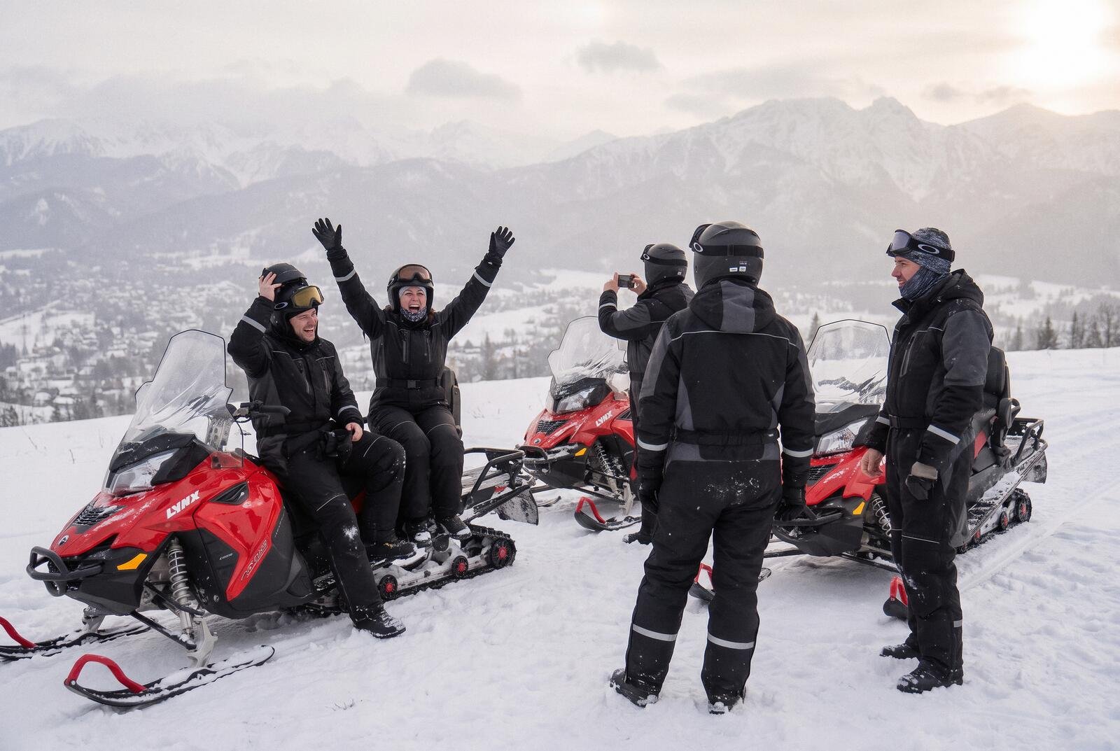 Para w kaskach z rękami w górze na skuterze śnieżnym na punkcie widokowym, obok reszta grupy, w dole Zakopane, na horyzoncie Tatry